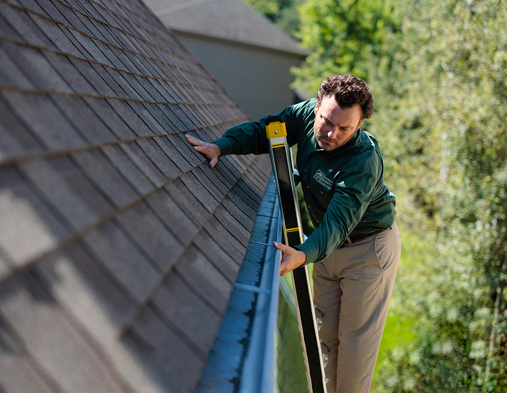 An NPI inspector stands on a ladder and examines the house's gutters.