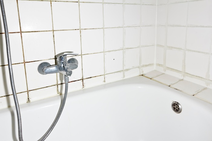 A bathroom wall with dirty grout above a bathtub with silver tubing.